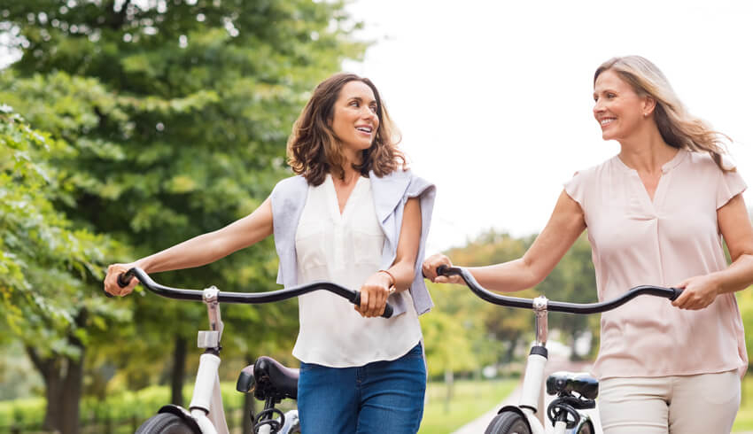 Two women with radiant skin walk their bikes along a park path 